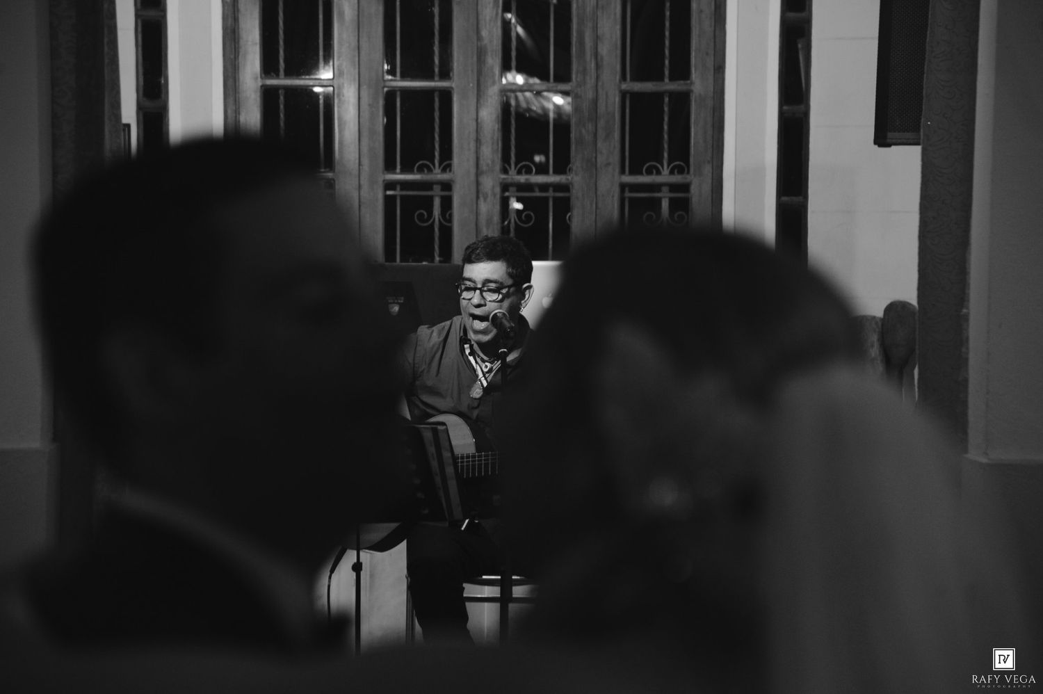 Black and white wedding photo of an elderly guest seated in a chair, captured between blurred foreground figures.