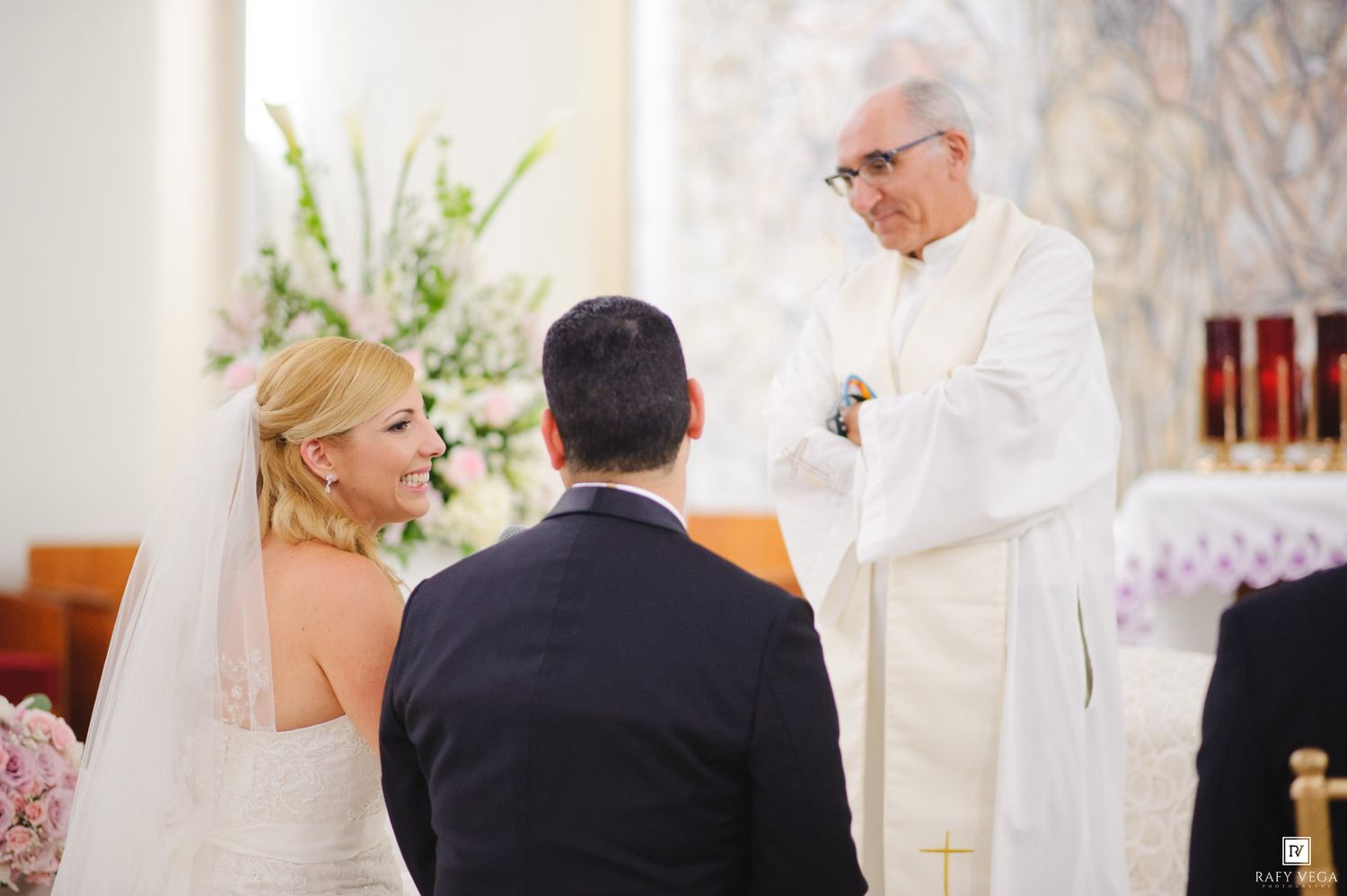 Bride and groom exchange vows before a priest at a church wedding ceremony with floral arrangements.
