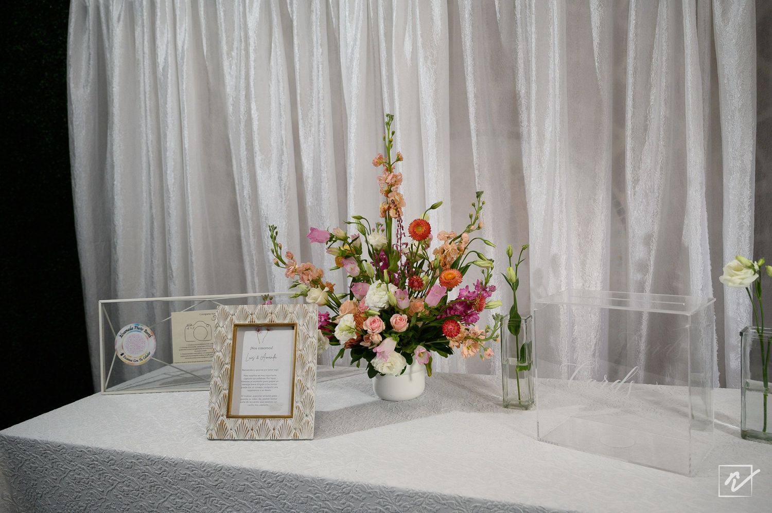 Floral arrangement of orange, pink and white blooms beside a framed sign and clear acrylic boxes on a white draped table at an event.