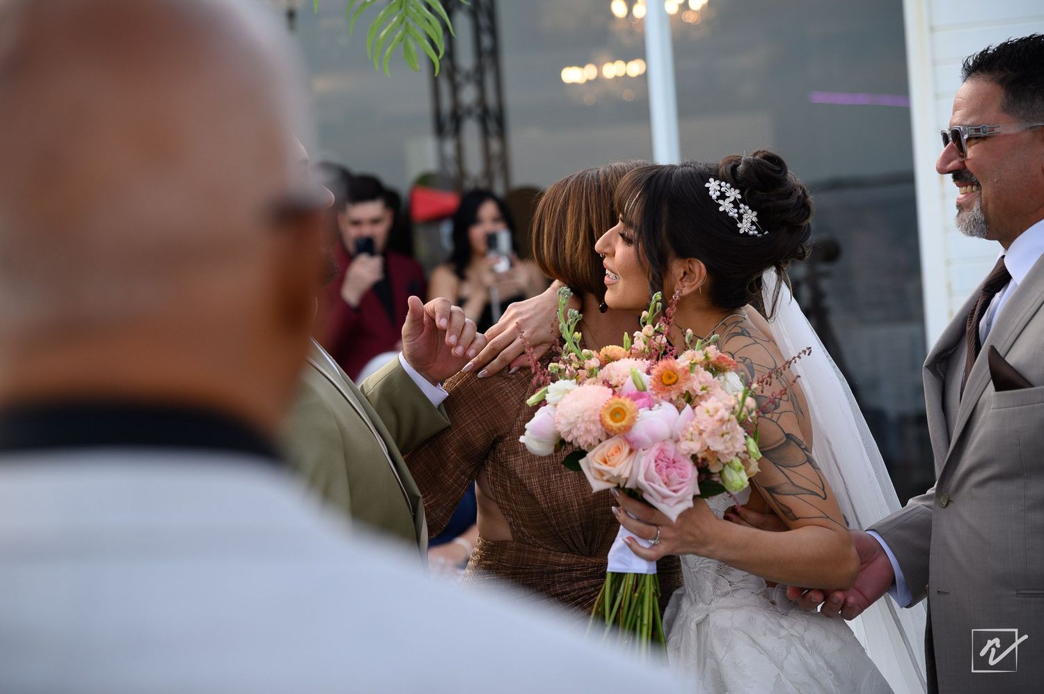 A bride in a white lace gown holding a pink and peach bouquet is embraced by a smiling man in a gray suit at a wedding ceremony.