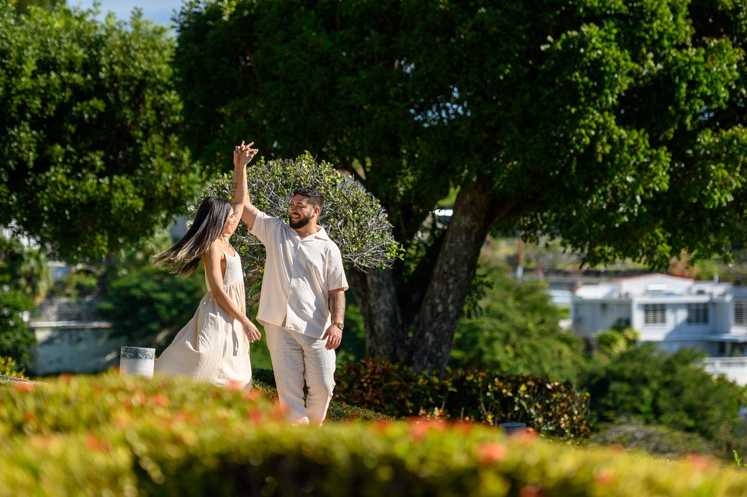A couple dances joyfully in a lush garden park with a large tree and colorful flowers in the background.