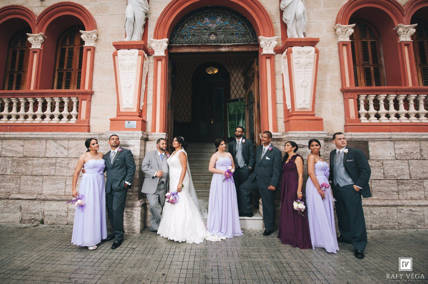 Wedding party poses in lavender gowns and gray suits before an ornate red building entrance.