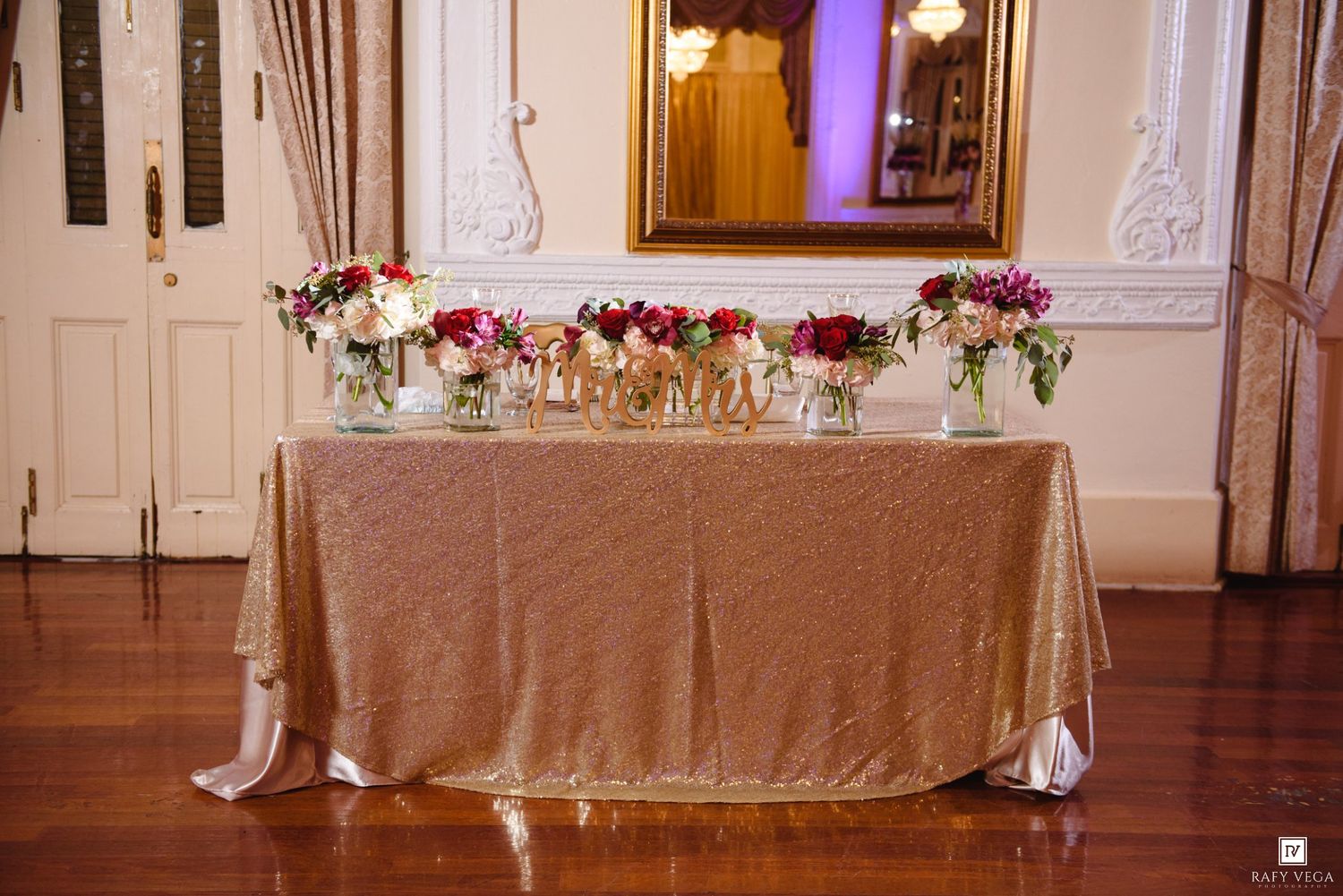 Elegant wedding sweetheart table with gold sequin tablecloth, floral centerpieces, and decorative gold letters in a grand ballroom.