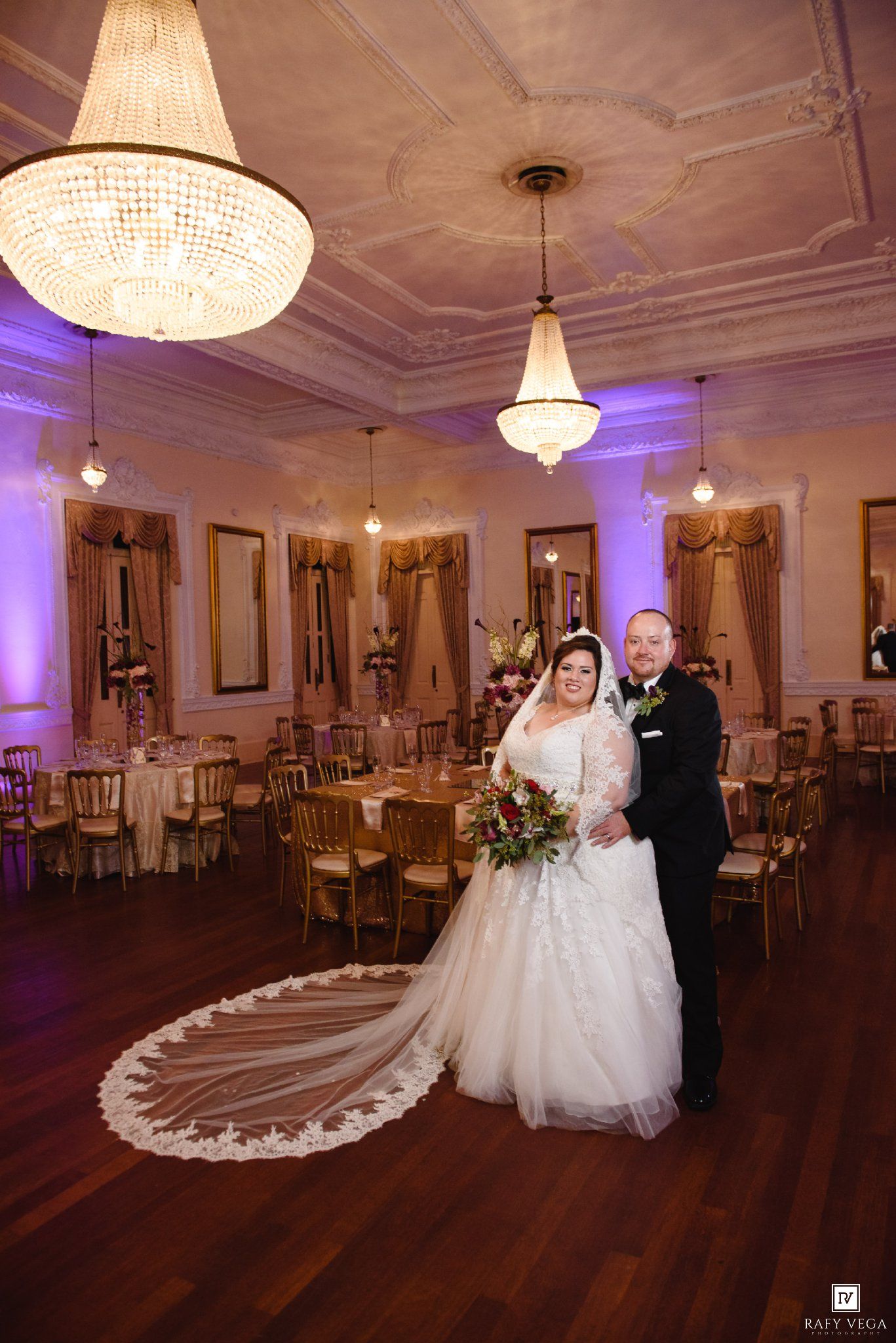 Bride and groom pose in elegant ballroom with crystal chandeliers, purple uplighting, and gold chiavari chairs.