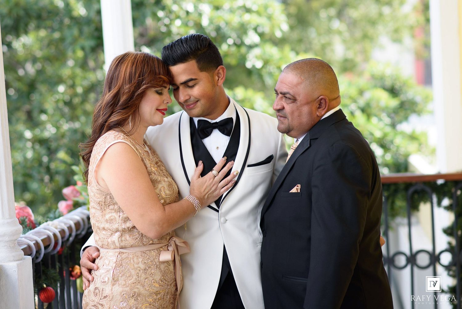 Groom in white tuxedo shares tender moment with parents at elegant outdoor wedding venue.