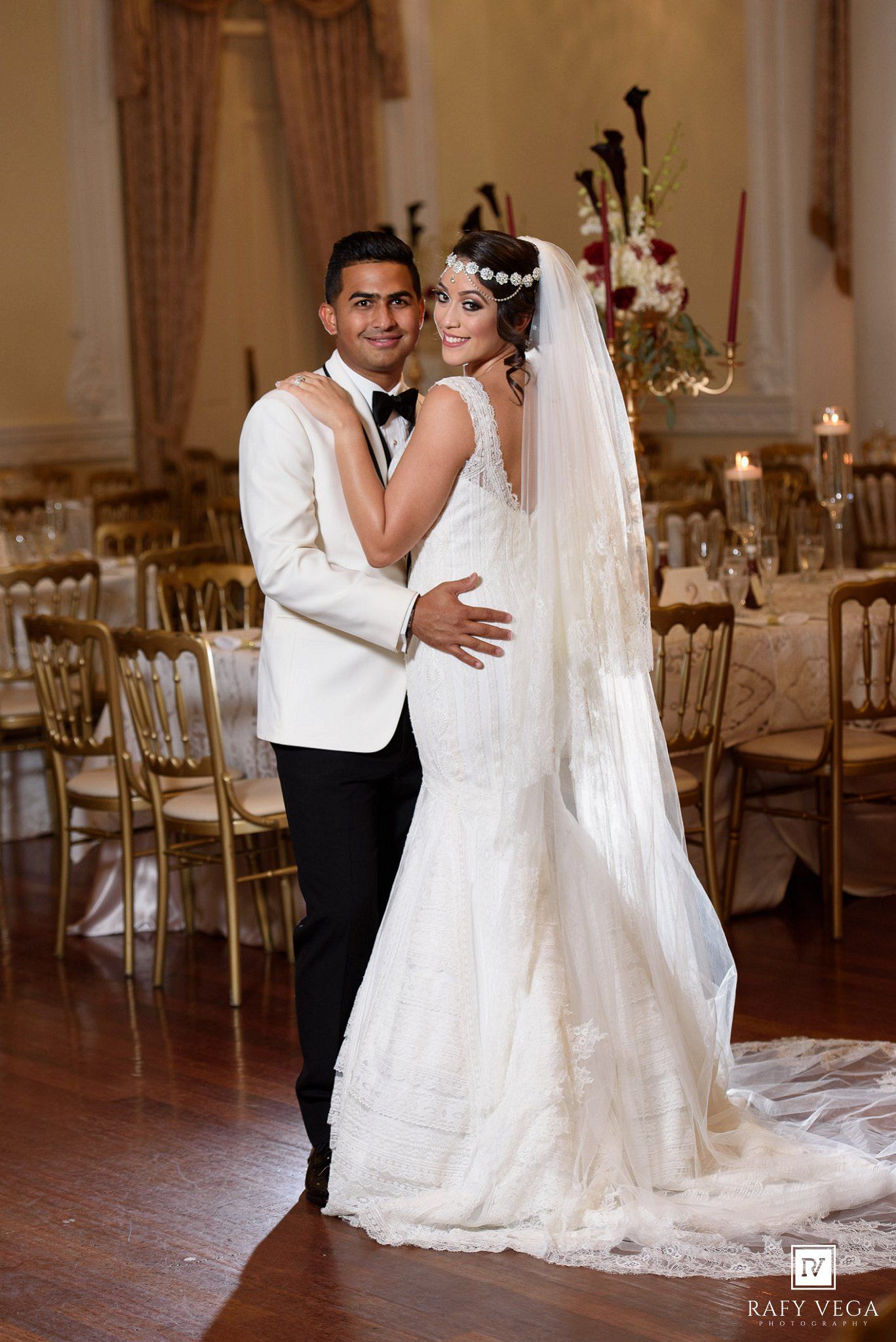 Bride in white mermaid gown and groom in white tuxedo pose elegantly in decorated wedding reception ballroom.