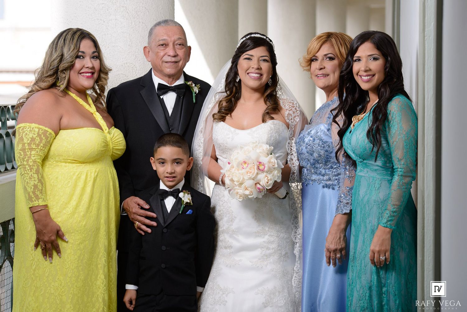 Bride in white gown poses with family including a young boy in tuxedo and women in colorful dresses at a wedding.