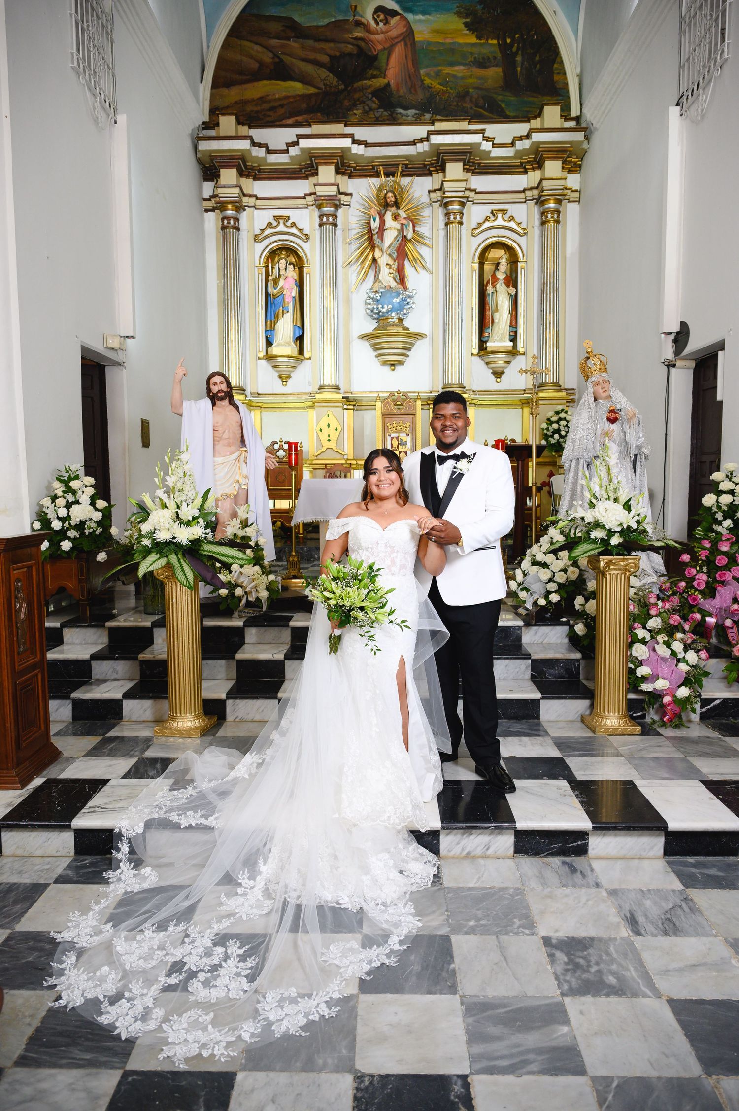 Bride and groom pose at ornate Catholic church altar with floral arrangements, bride in white gown with lace veil, groom in tuxedo.