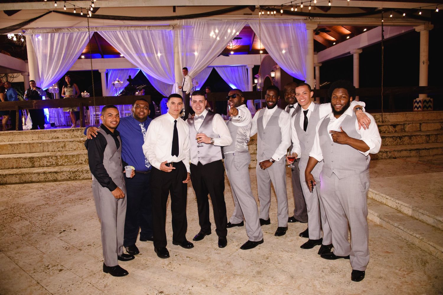 Groom poses with groomsmen in gray suits at a beautifully decorated outdoor wedding reception venue.