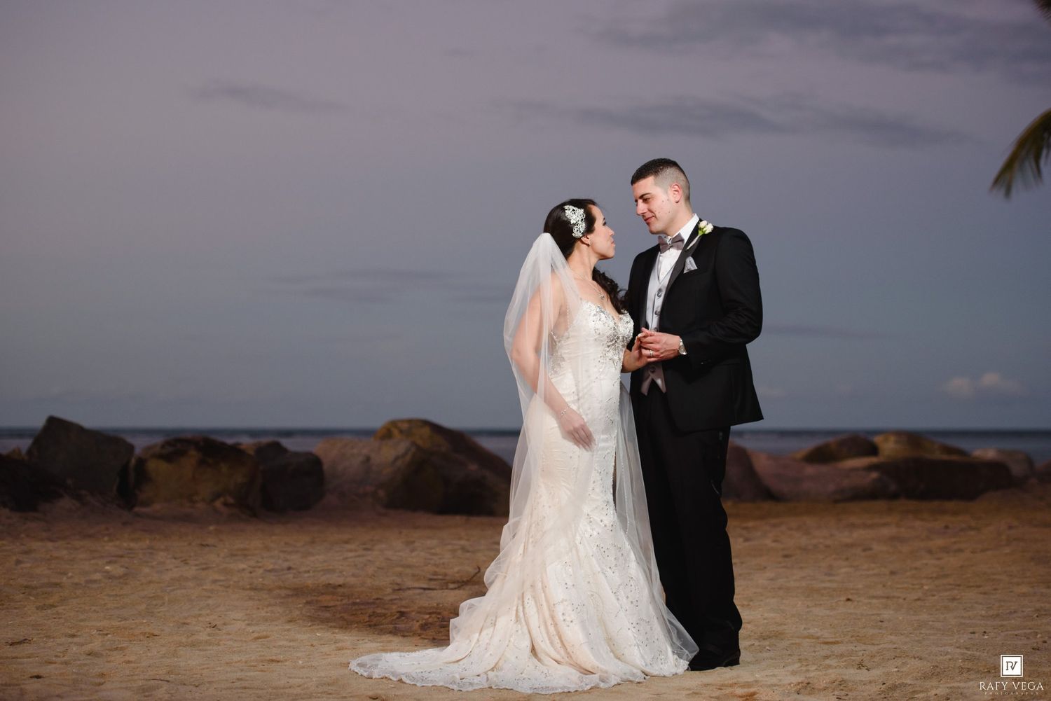 Bride and groom share a romantic moment on a sandy beach at dusk, with rocks and ocean in the background.