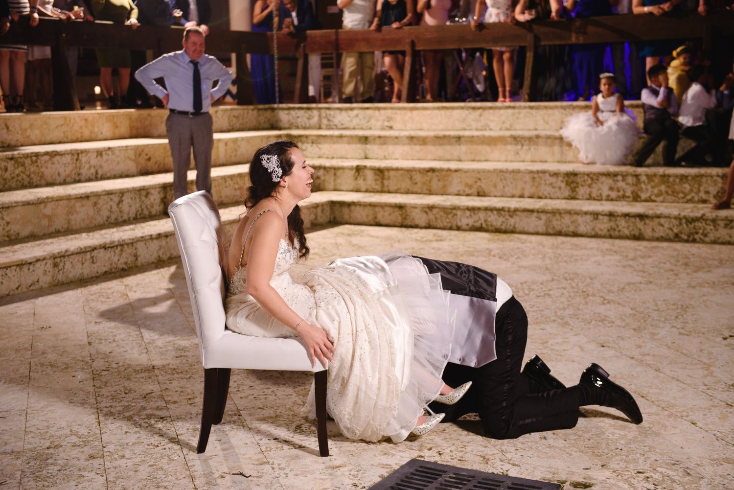 Groom kneeling before bride seated on white chair at elegant outdoor wedding reception.