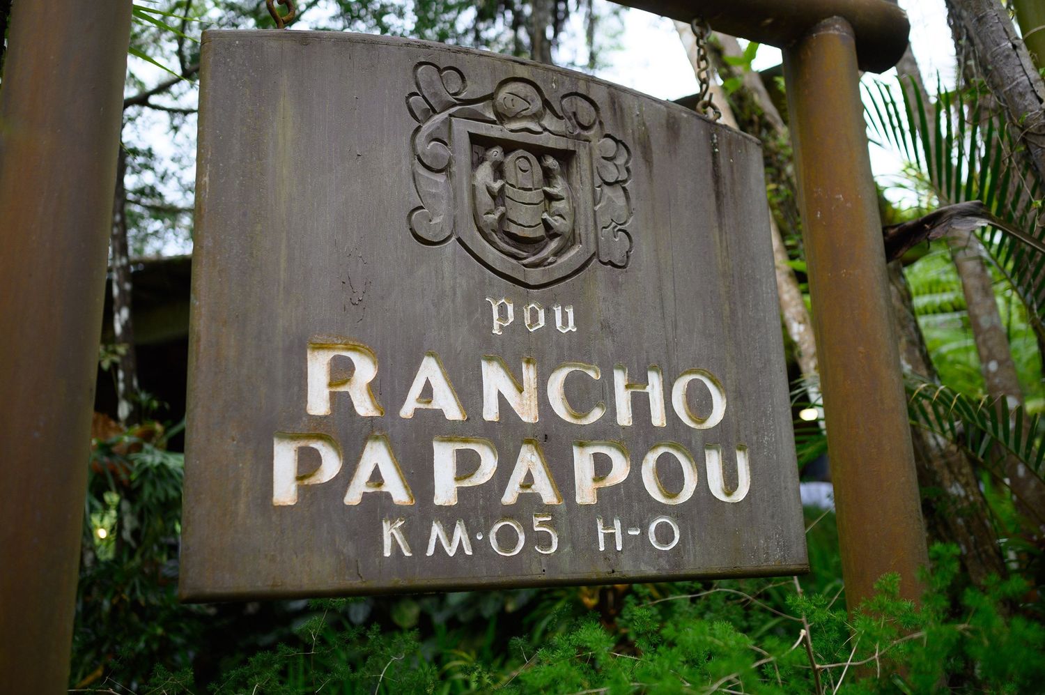 Wooden sign for Rancho Papapou KM.05 H.O with a coat of arms emblem, surrounded by lush green tropical foliage.