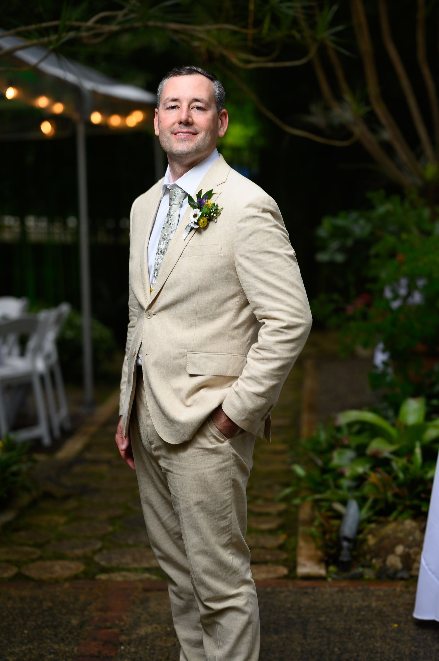 Man in beige linen suit with boutonniere smiling in garden wedding venue with string lights.