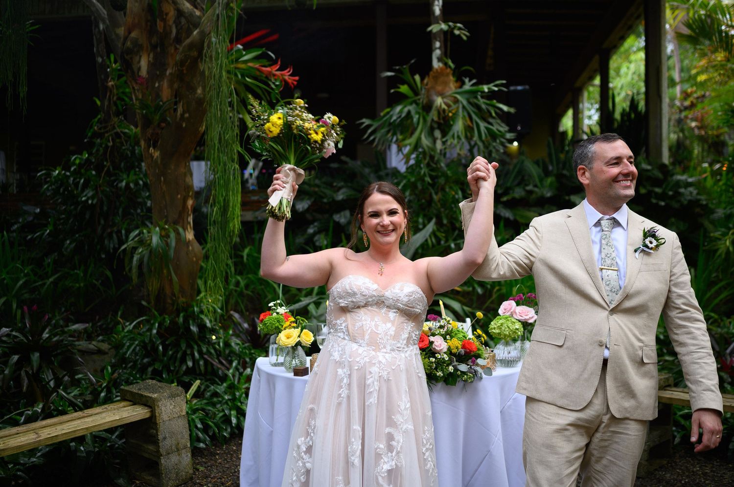 Joyful newlyweds celebrate outdoors in lush tropical garden, bride in lace gown holding colorful bouquet aloft.
