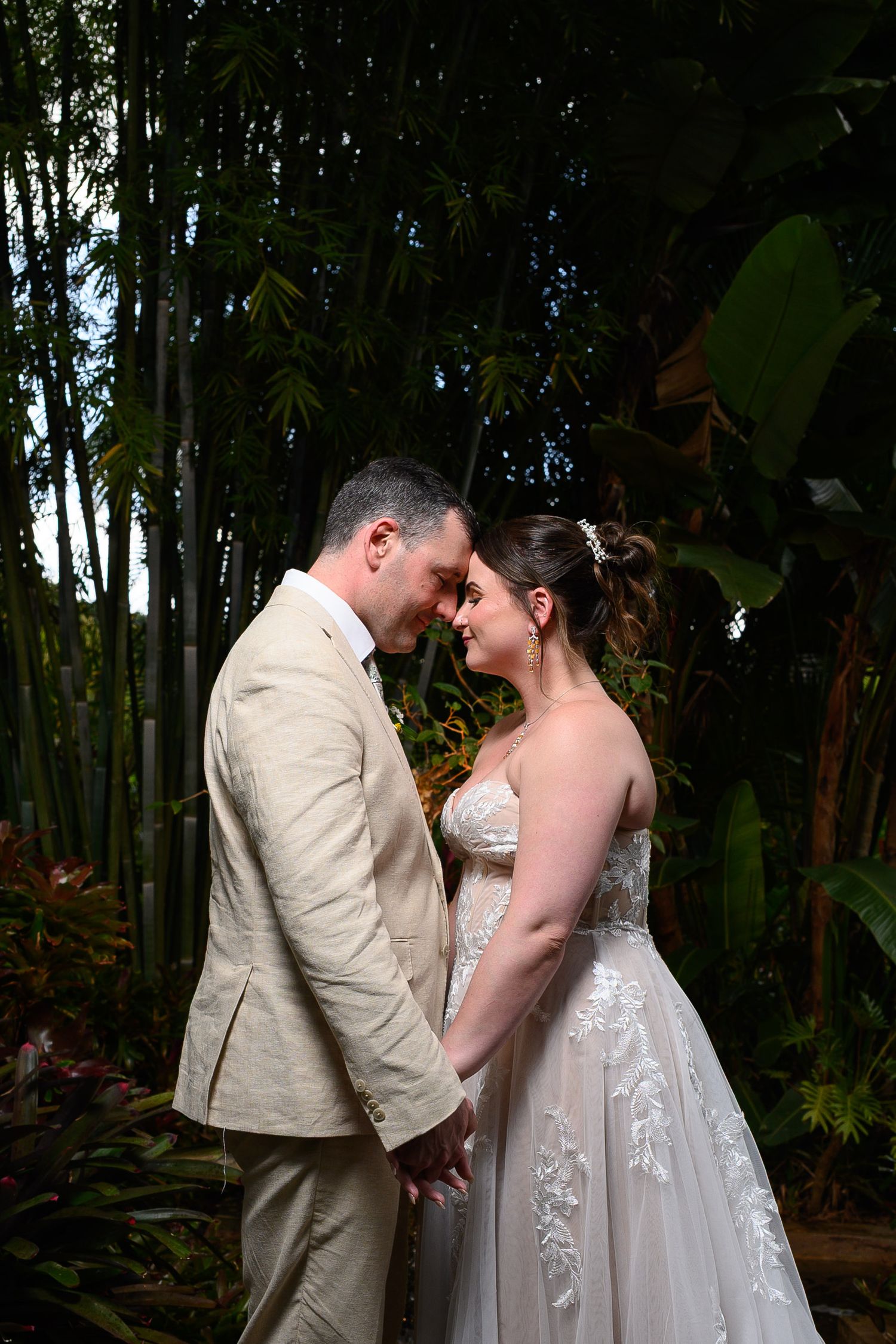 Bride and groom holding hands forehead to forehead in lush tropical garden, groom in tan suit, bride in lace wedding gown.