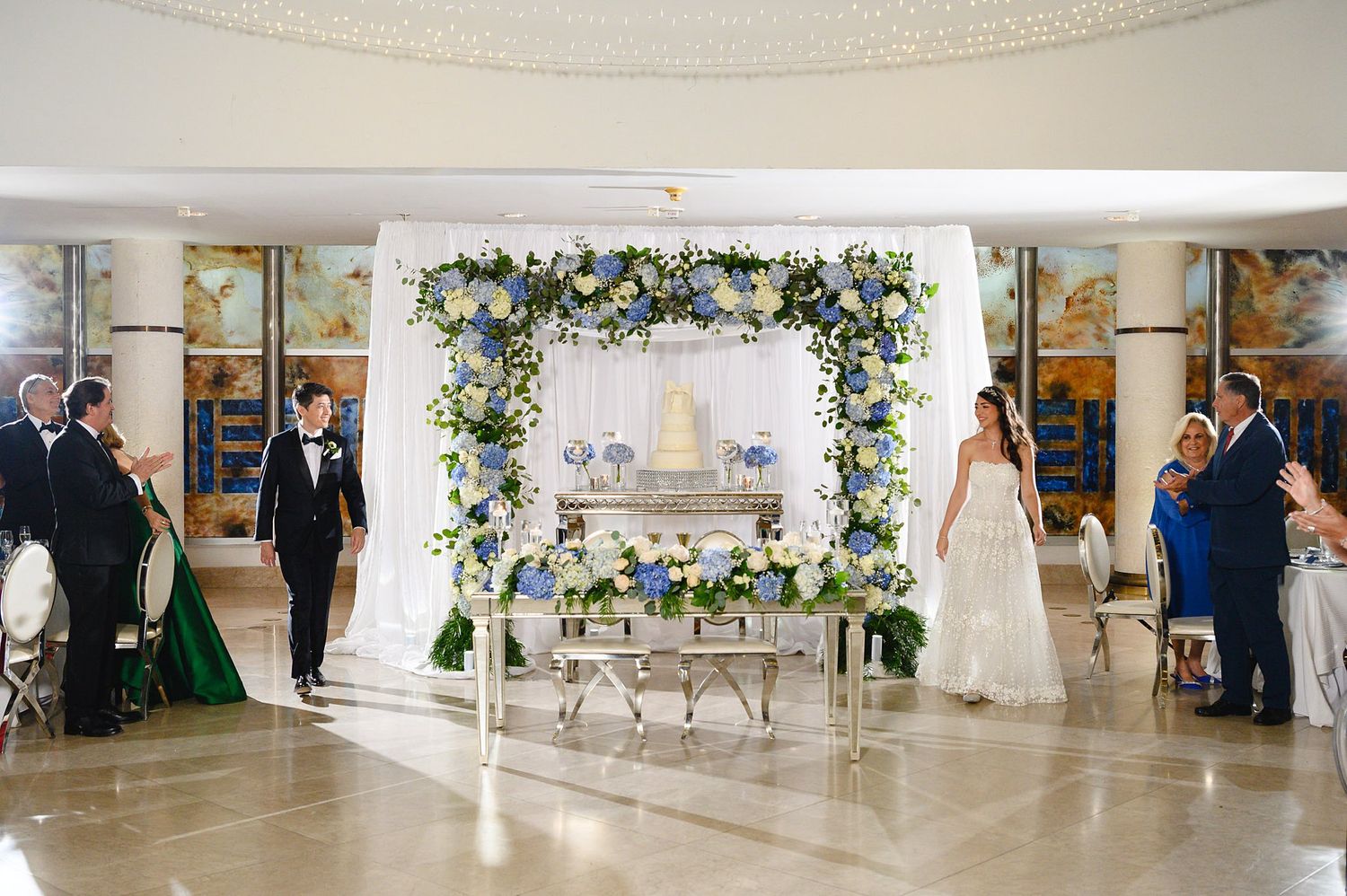 Elegant wedding ceremony with floral arch backdrop, white draping, and blue and white flowers in a grand ballroom venue.