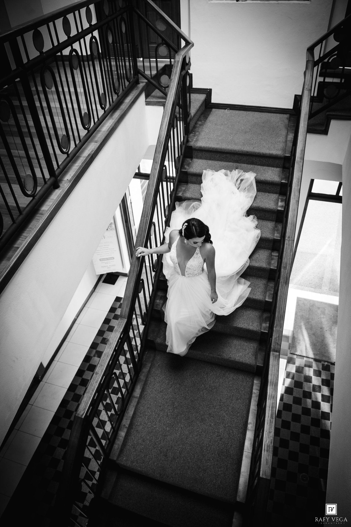 Bride in white wedding gown descending elegant staircase, captured from above in black and white photography.
