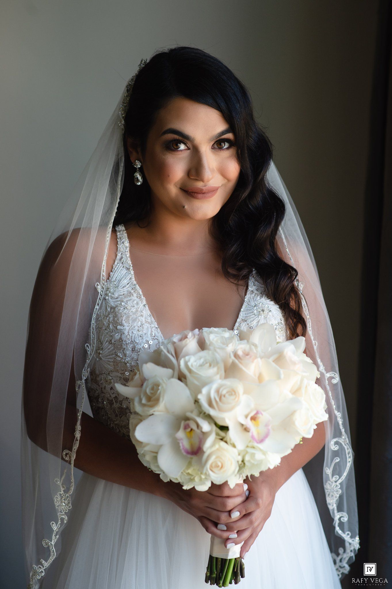 Bride in beaded V-neck gown and veil holds white rose and orchid bouquet, smiling softly in elegant bridal portrait.