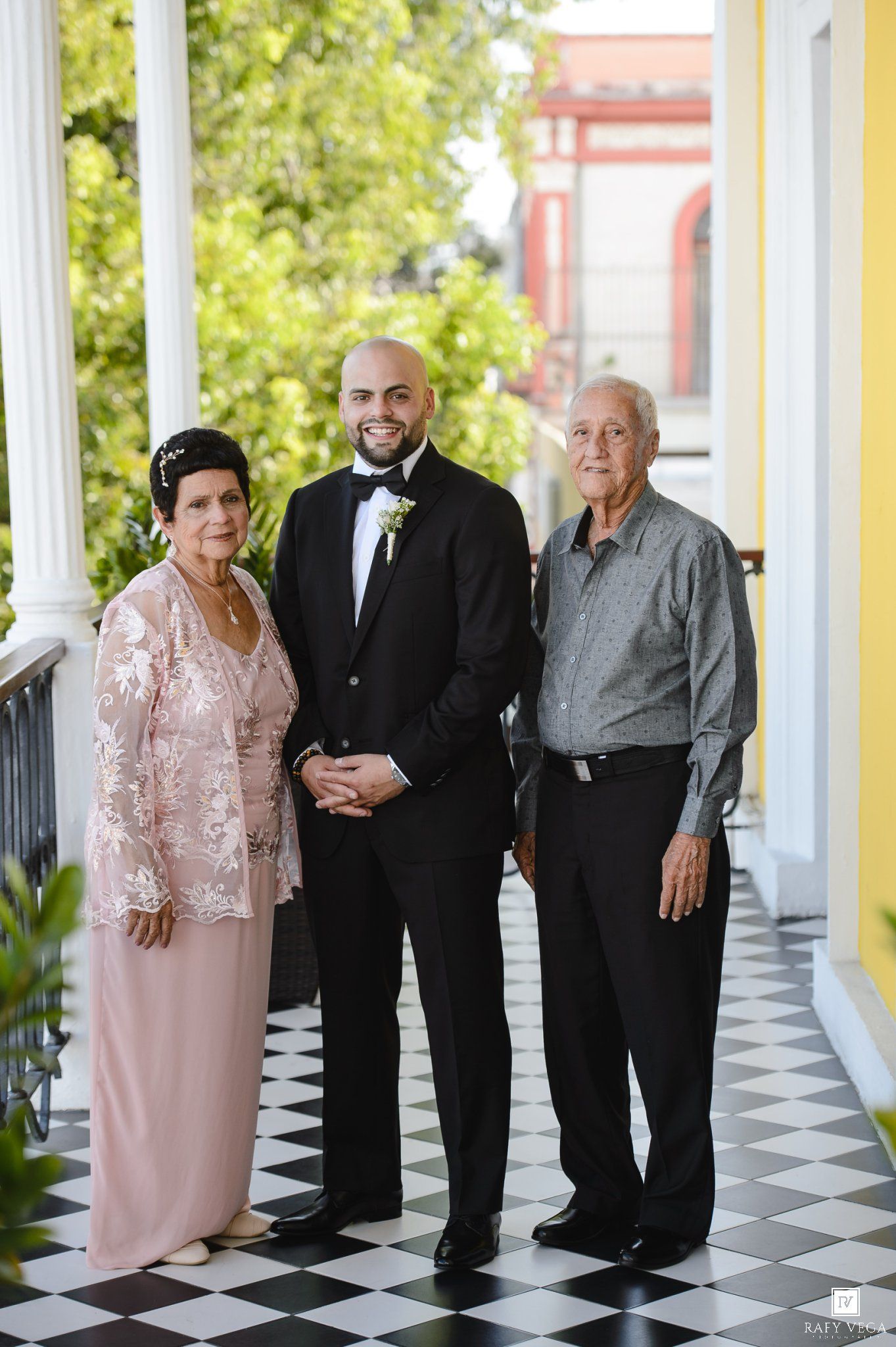 Groom in black tuxedo poses with parents on checkered porch, mother in pink lace gown at wedding venue.