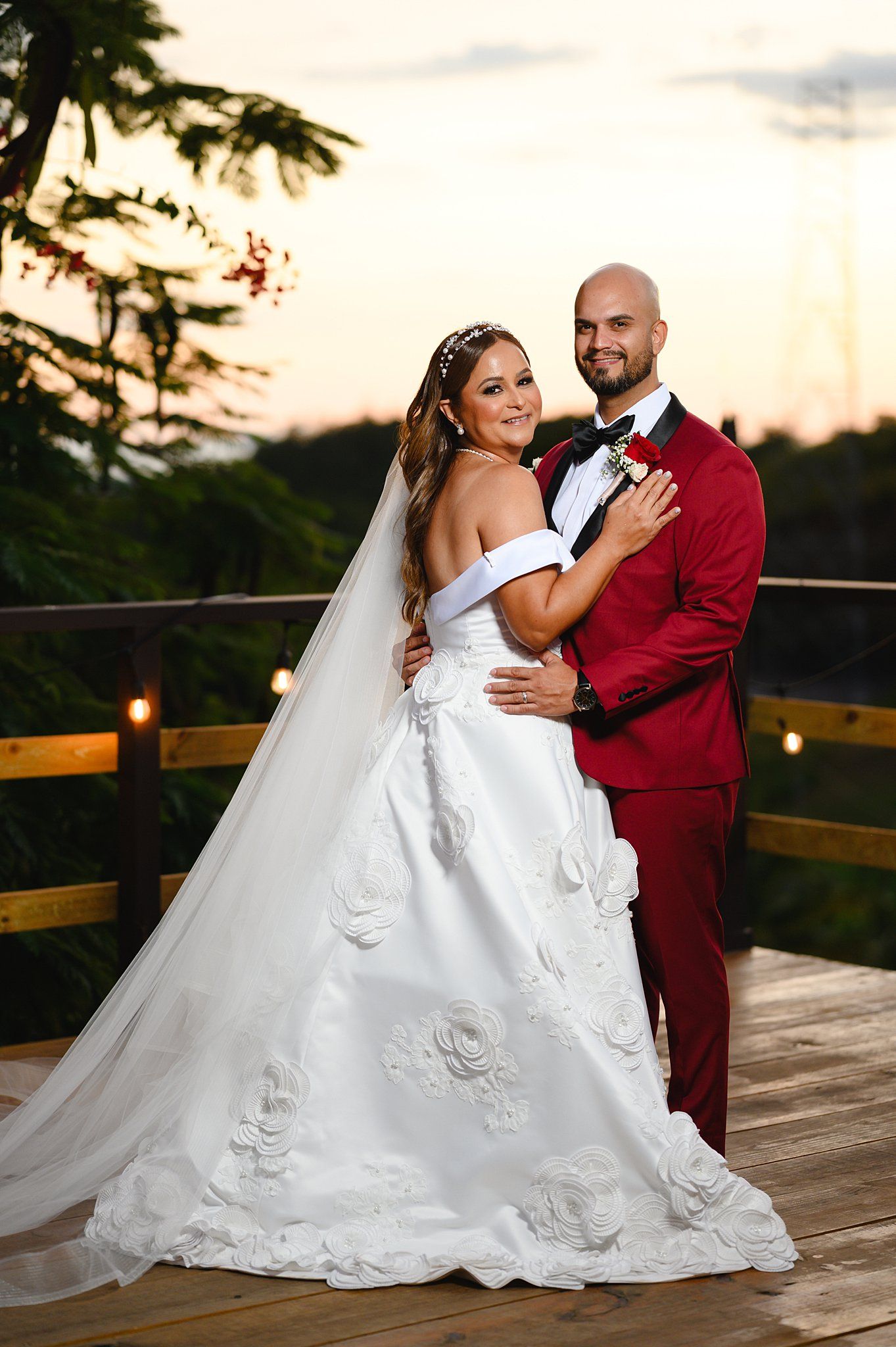 Bride in white floral ball gown and groom in red suit pose romantically on wooden deck at sunset.