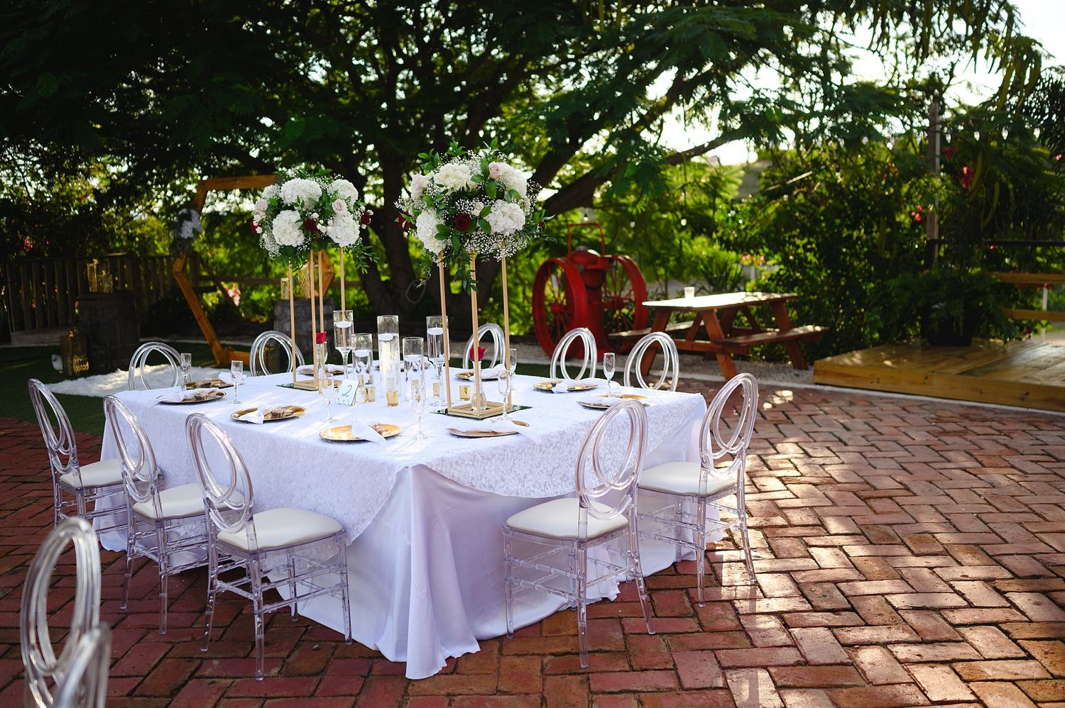 Elegant outdoor wedding reception table with white linens, floral centerpieces, and ghost chairs on a brick patio.