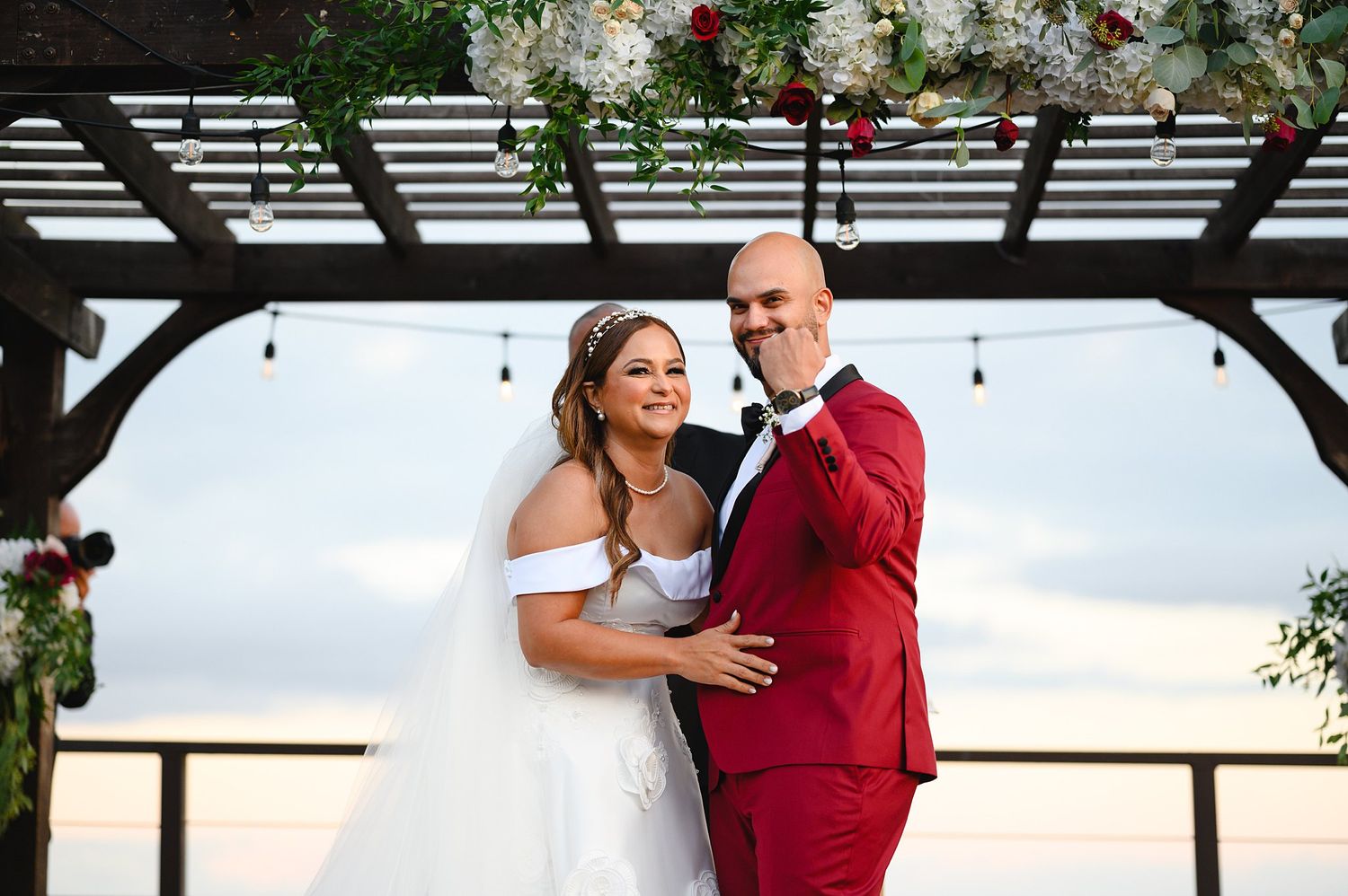 Smiling bride and groom in red suit pose together under a floral-adorned pergola with string lights at twilight.