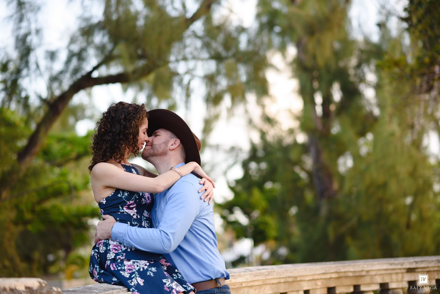 A couple shares a romantic kiss outdoors, surrounded by lush green trees in a park setting.