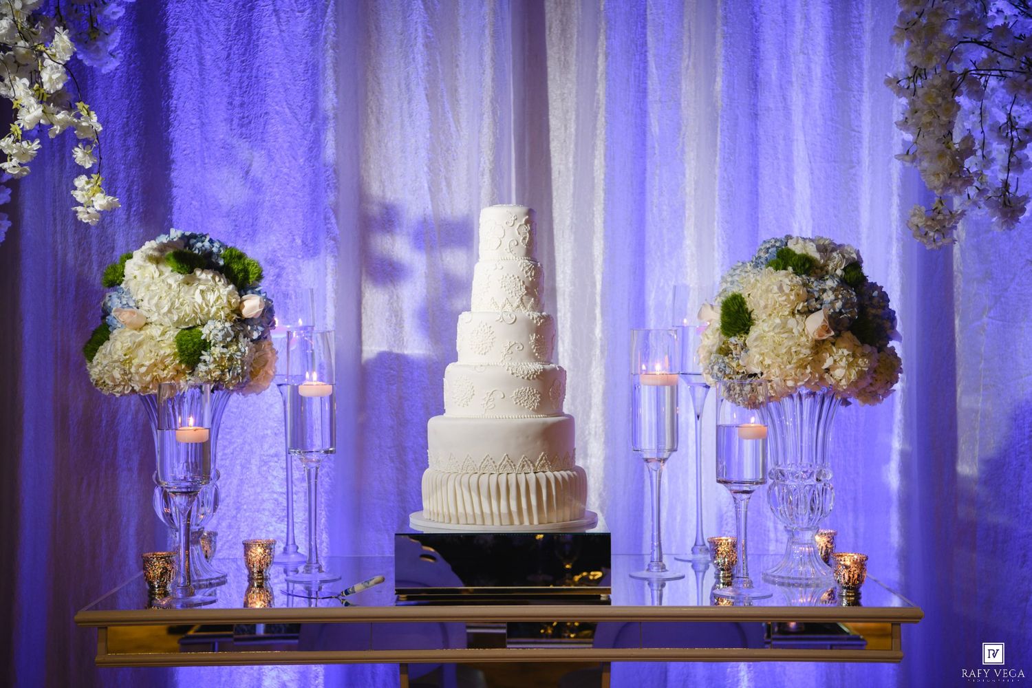 Elegant five-tier white wedding cake on a gold table with floral arrangements and candles against a purple-lit backdrop.
