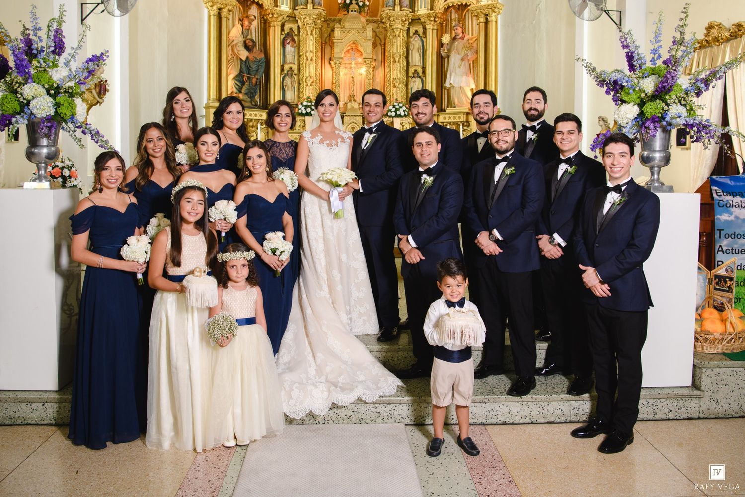 Wedding party poses in church with bride and groom, bridesmaids in navy blue gowns, groomsmen in suits, and flower girls.