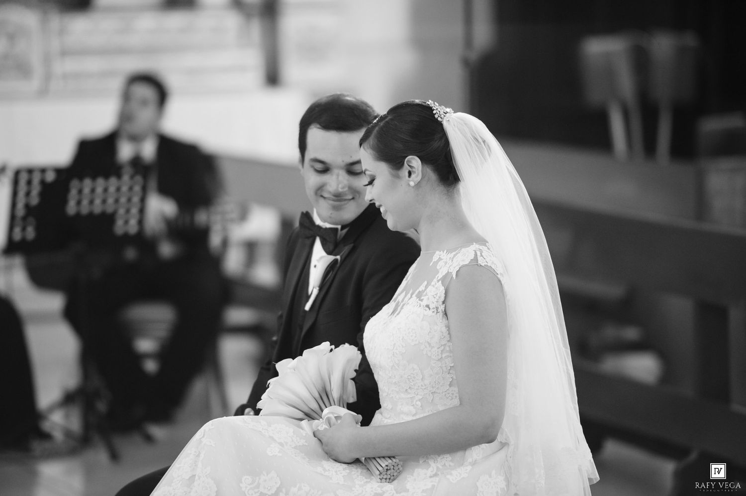 Black and white photo of smiling bride and groom sitting together during wedding ceremony in church.