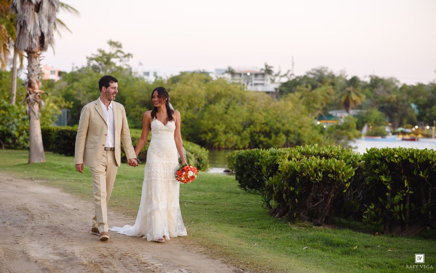 Newlywed couple walks hand-in-hand along scenic tropical path at dusk with bouquet.