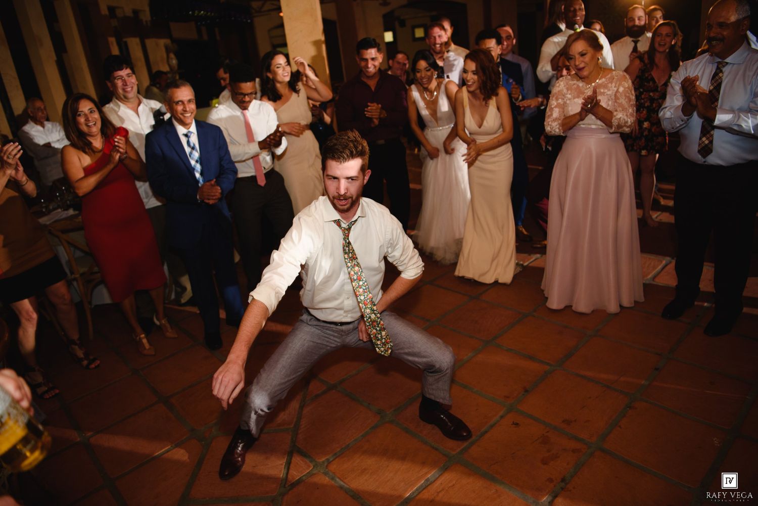 Man performing energetic dance moves at wedding reception while guests clap and cheer in background.