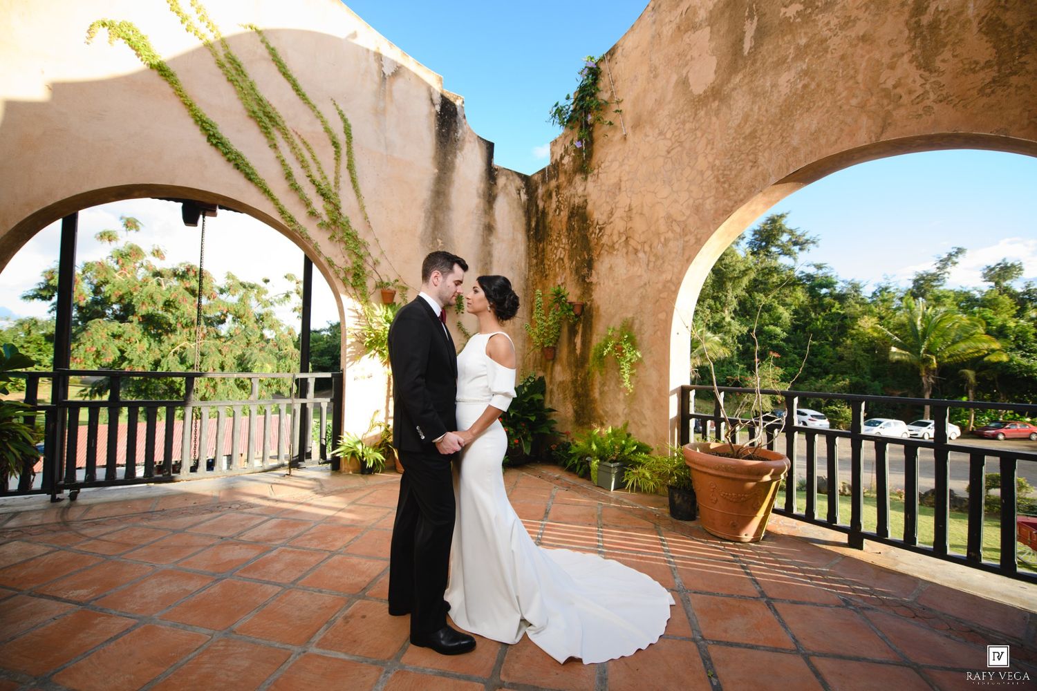 Bride and groom share a kiss on a terracotta patio with arched walls and tropical gardens.