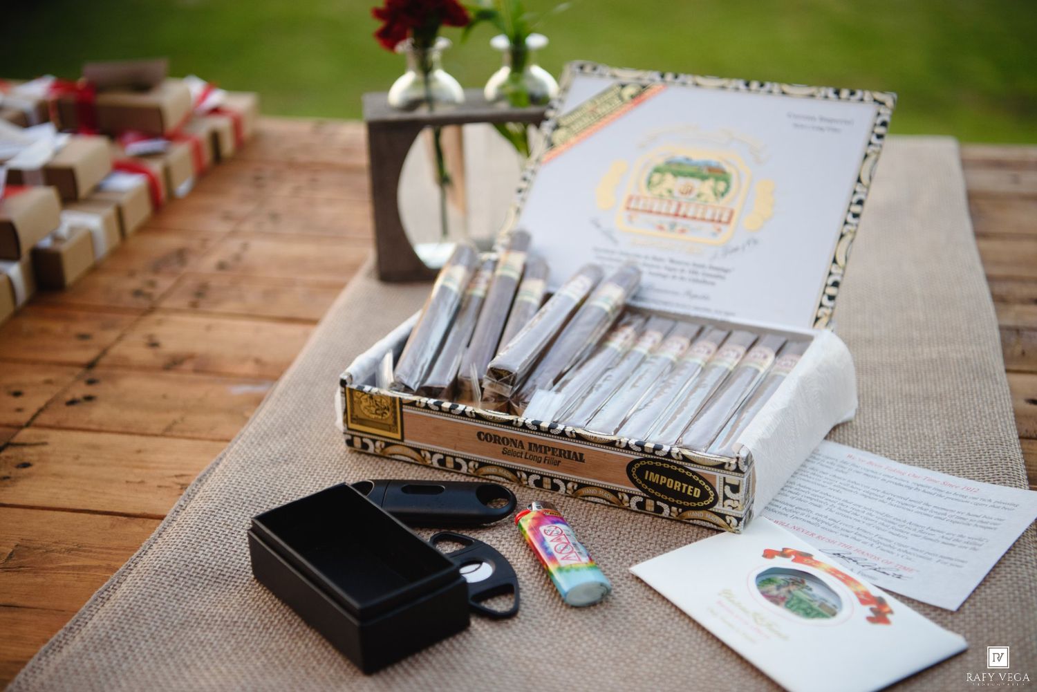 Open cigar box with wrapped cigars displayed on rustic wooden table at outdoor wedding reception.