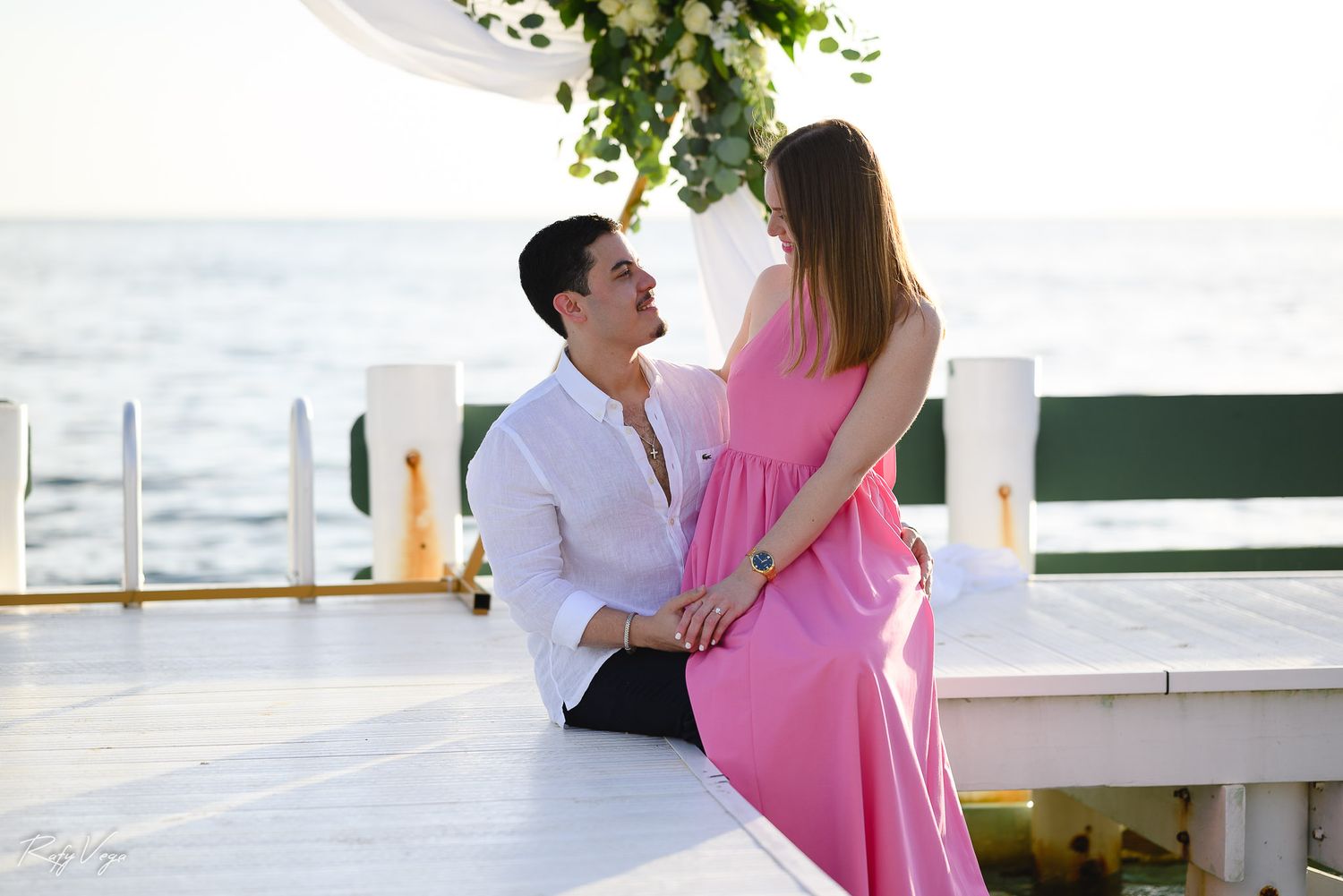 Man kneeling before woman in pink dress during romantic seaside proposal on white deck with floral arch.