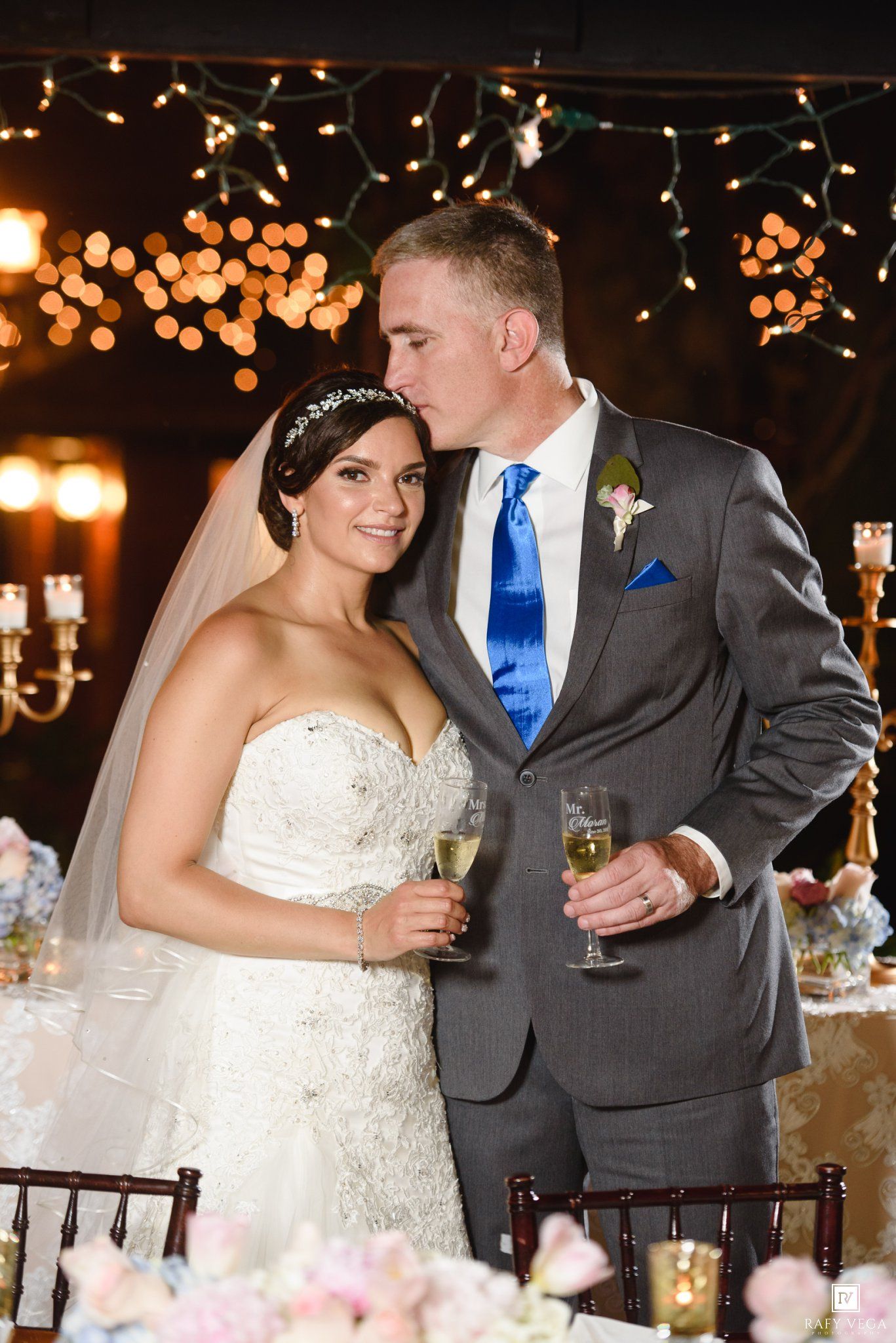 Bride and groom share a romantic toast with champagne at their elegant wedding reception with warm string lights.