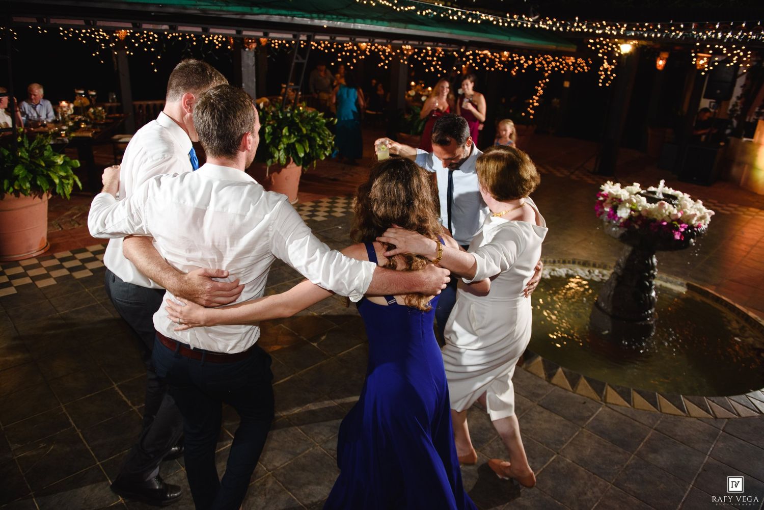 Wedding guests form a circle dance on a checkered outdoor dance floor at a nighttime reception.