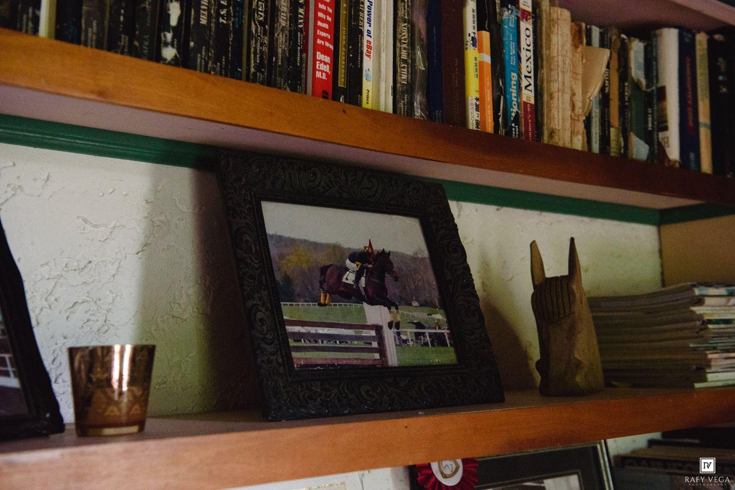Framed horse jumping photo displayed on wooden bookshelf with books and decorative items.