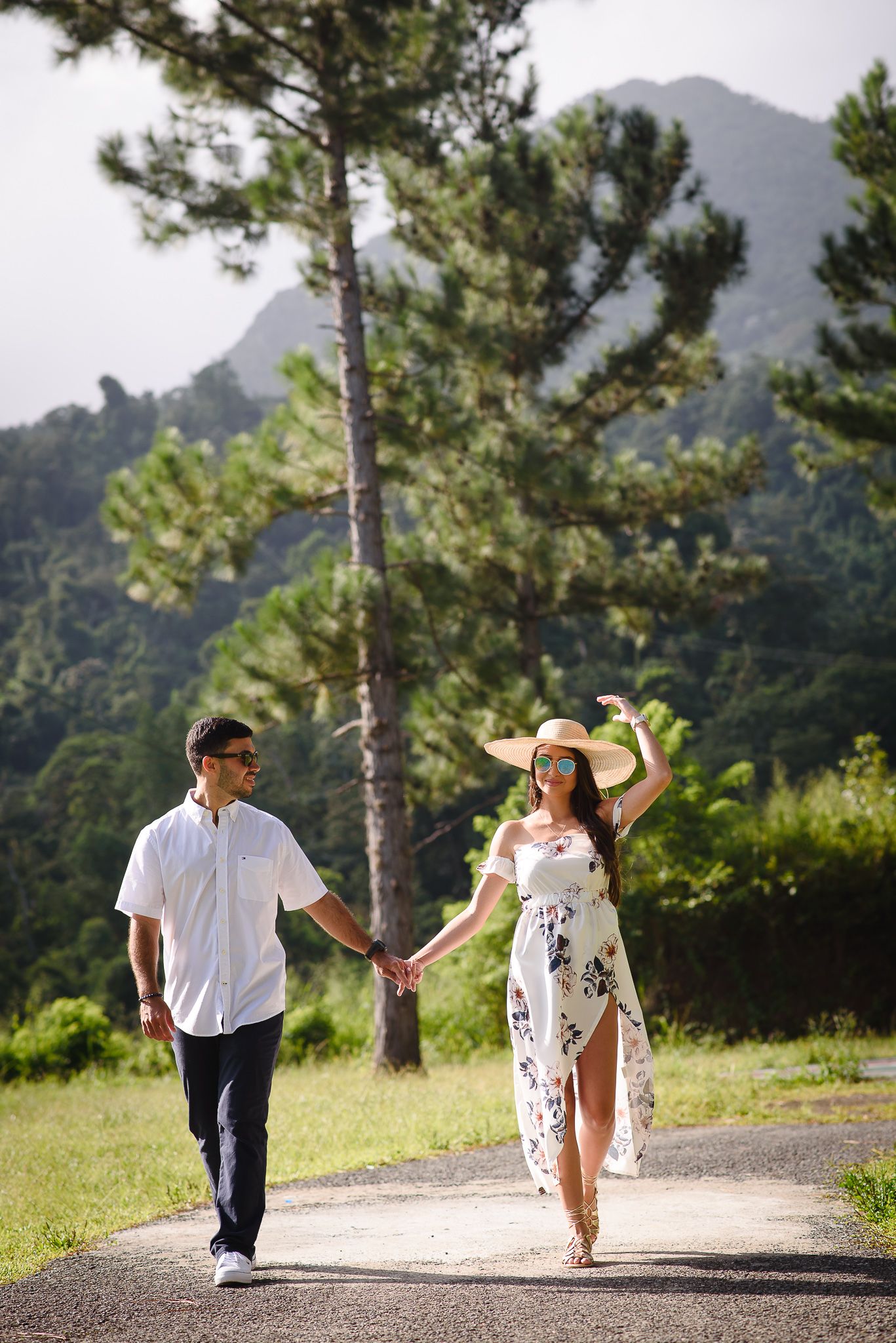 Couple holding hands walking on a path surrounded by lush green trees and mountains on a sunny day.