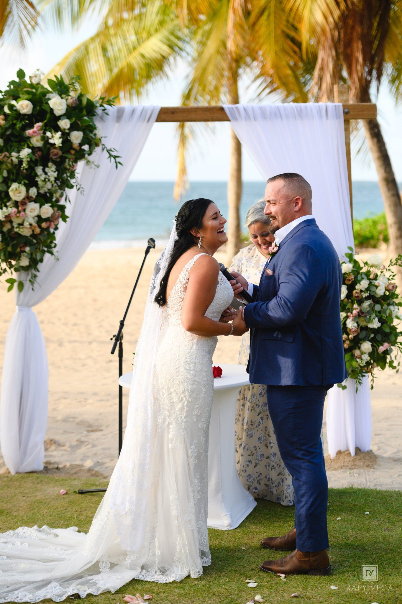 Couple exchanging vows at a beach wedding ceremony under a floral arch with white draping and palm trees.