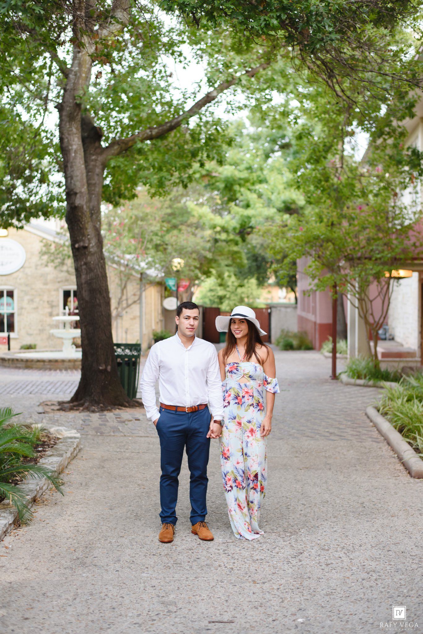 Couple holding hands on a tree-lined brick path, man in white shirt and woman in floral maxi dress with white hat.