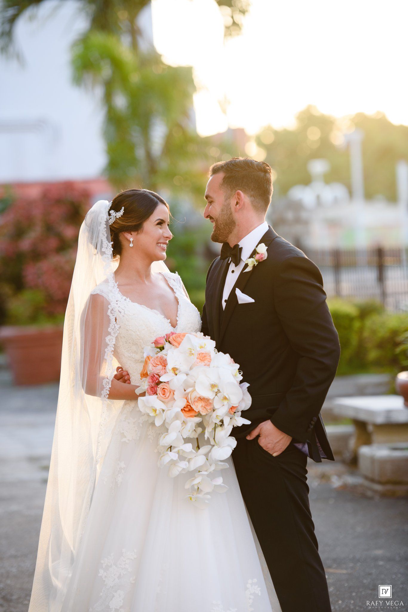 Bride and groom share a romantic glance outdoors at golden hour, she holds white orchid and peach rose bouquet.