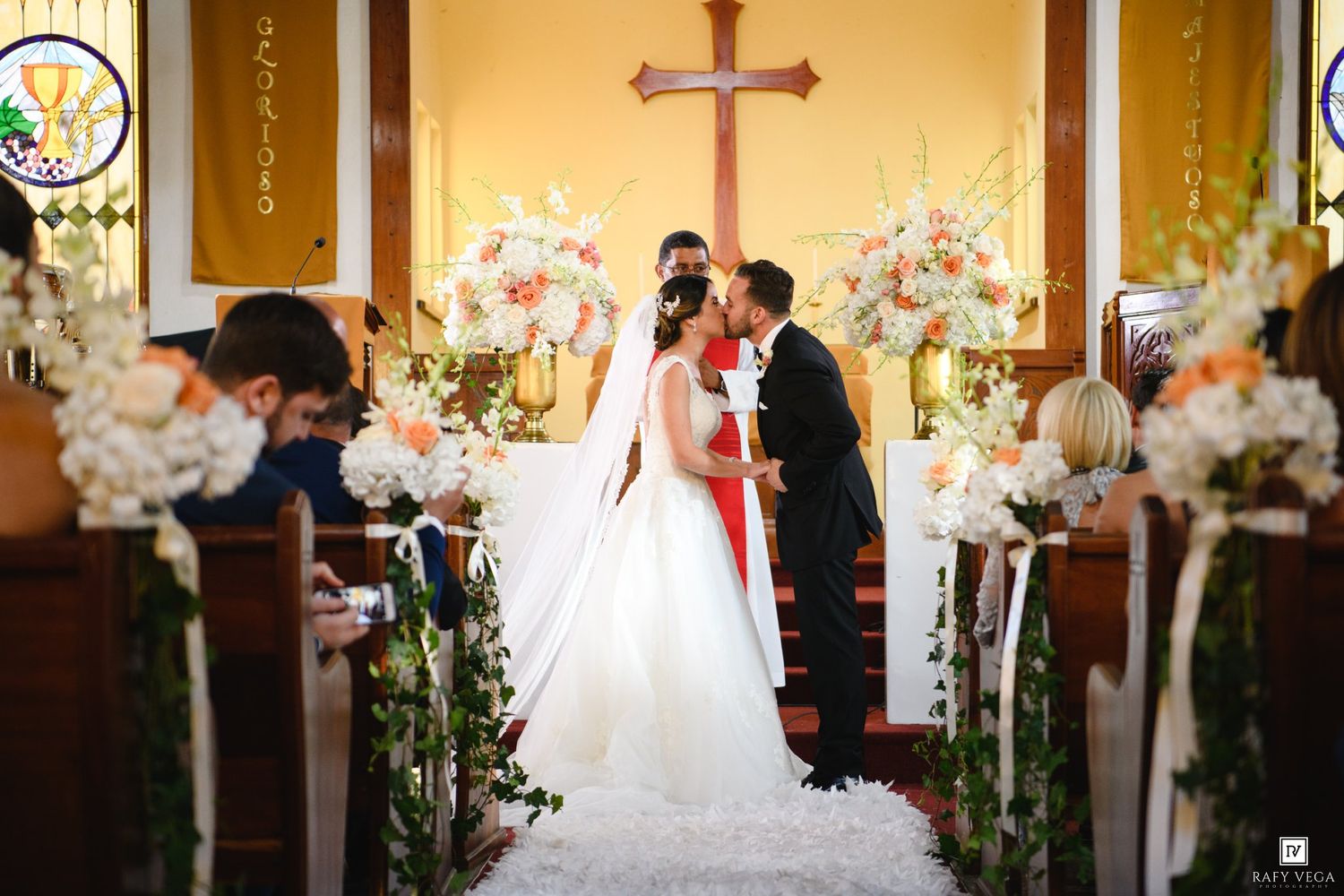 Bride and groom share a kiss at the altar of a church decorated with elegant white and peach floral arrangements.