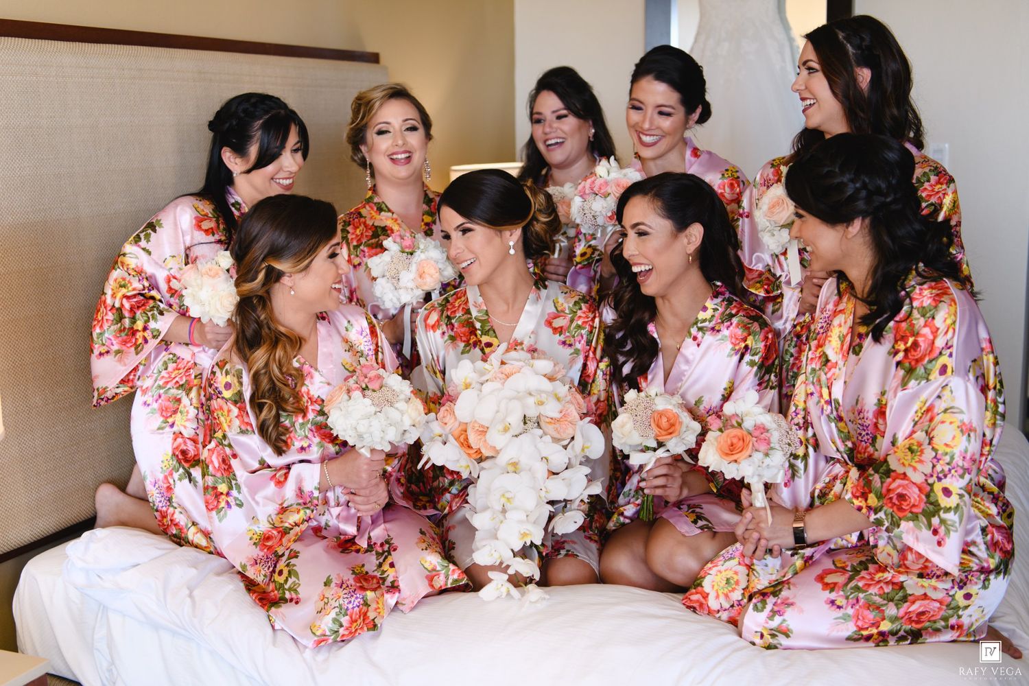 Bride and bridesmaids wearing matching floral robes sitting together on a bed holding white bouquets before the wedding.