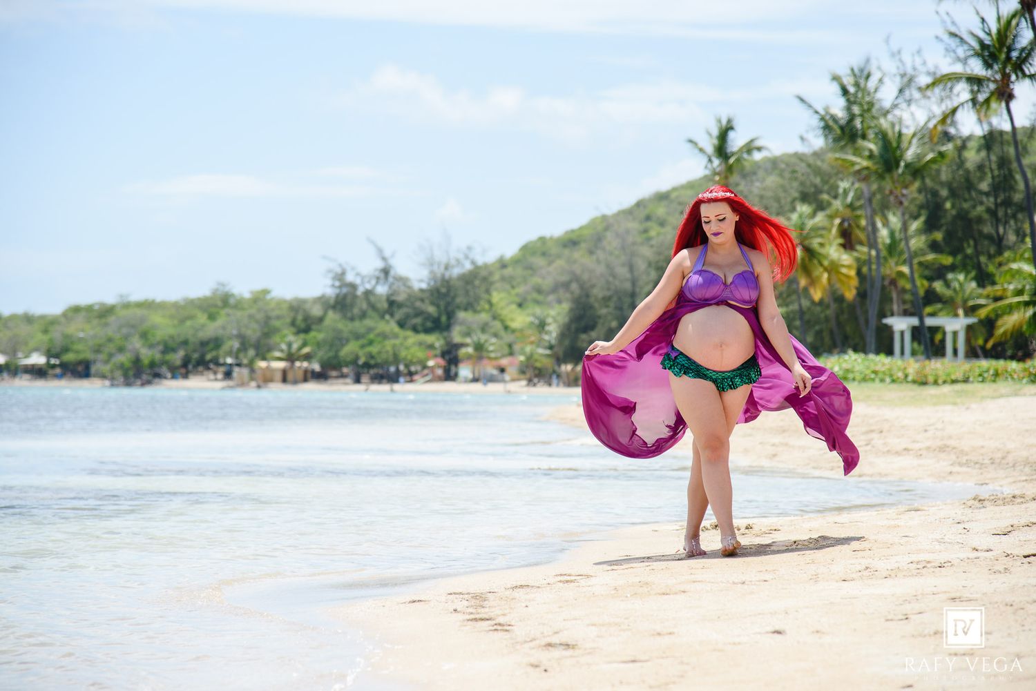 Woman in purple bikini with red hair walks on tropical beach holding flowing pink sarong with palm trees in background.
