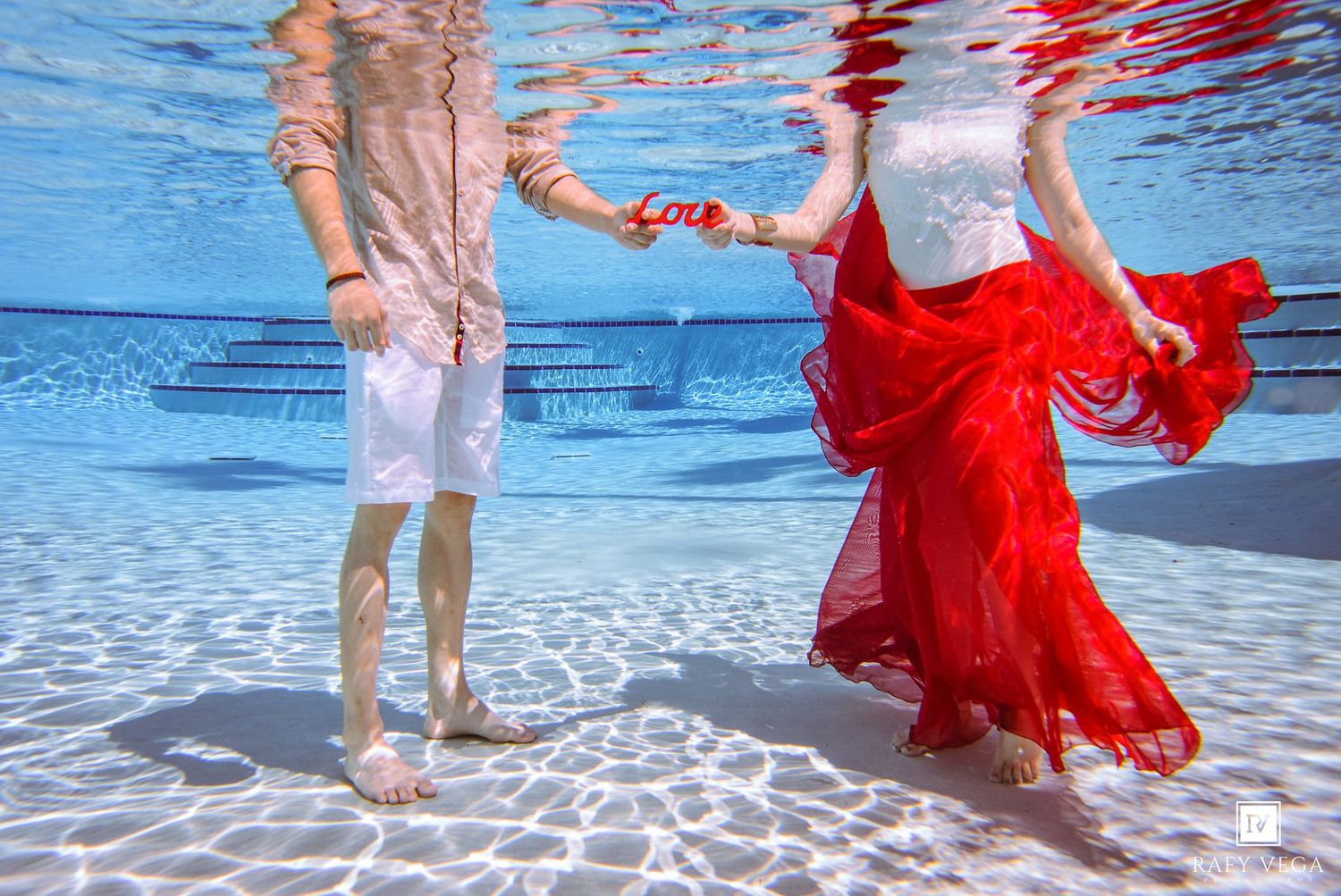 Couple holding hands underwater in a pool, woman in red flowing skirt and white top, man in white shorts.