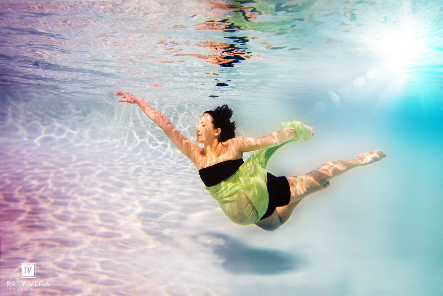 Woman in black swimsuit and green flowing fabric gracefully swimming underwater in a sunlit pool.