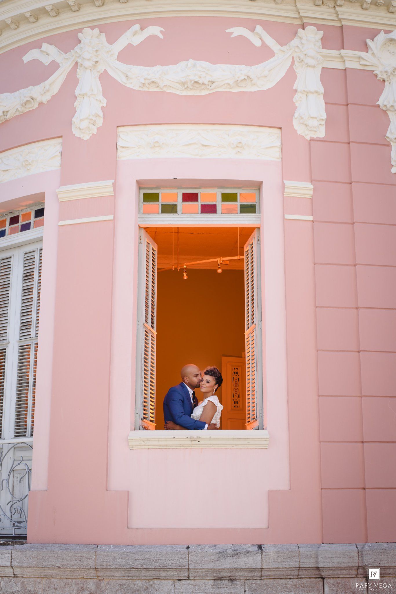 Wedding couple embracing in an ornate pink building window with colorful stained glass accents.