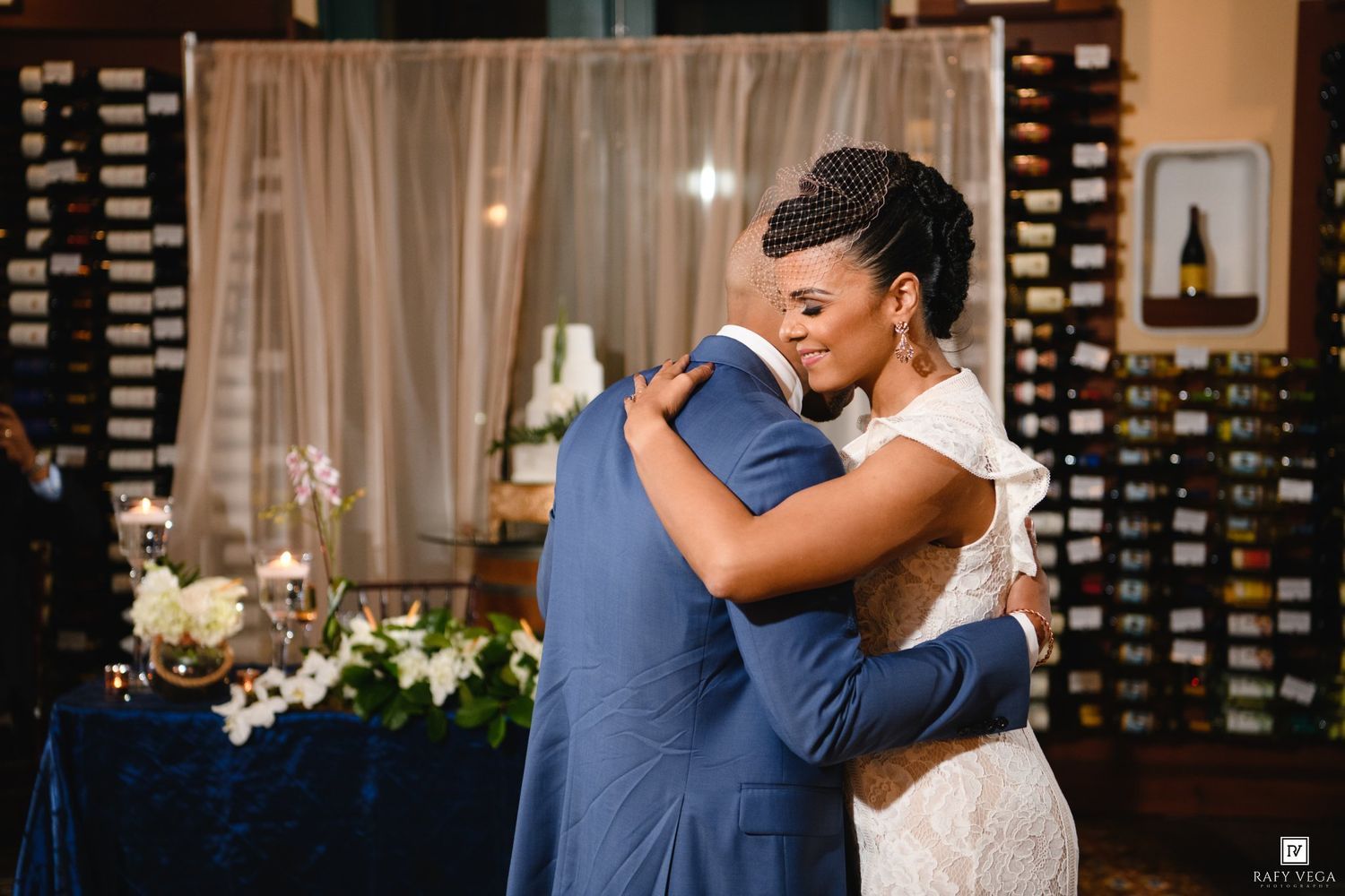 Bride and groom share an emotional first dance embrace at elegant wedding reception with wine wall backdrop.