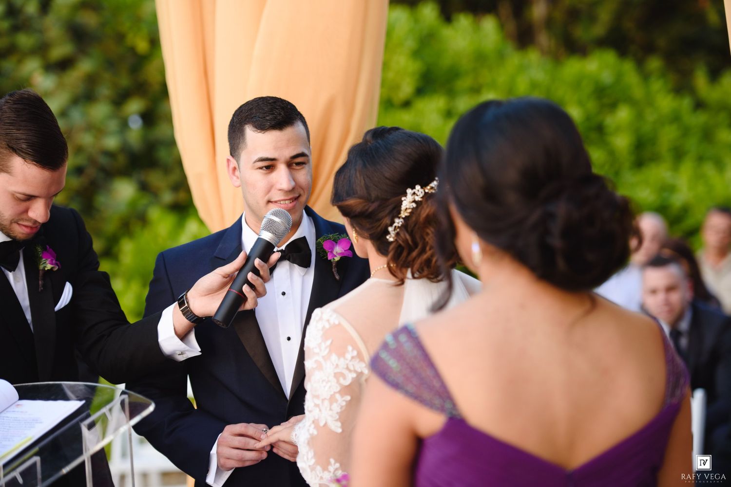 Groom reciting wedding vows into microphone at outdoor ceremony with bride and bridesmaid in purple dress.